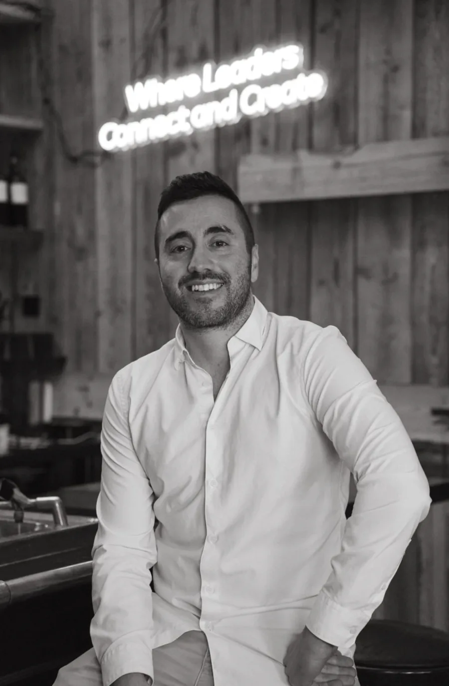 Alon Shkuri, producer, in black and white, smiling, at a bar with a neon sign behind him